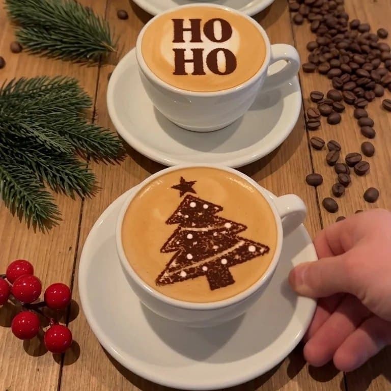 Two finished lattes, one dusted HO HO and one dusted with a Christmas tree. Example designs; included cards vary.